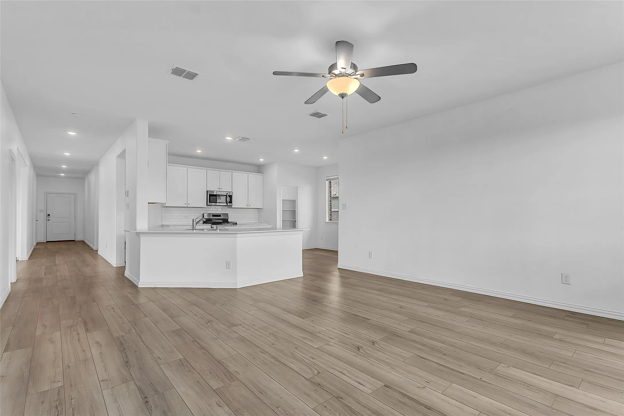 Open-concept living room flowing into the kitchen and front hallway, with a ceiling fan and wood-look flooring