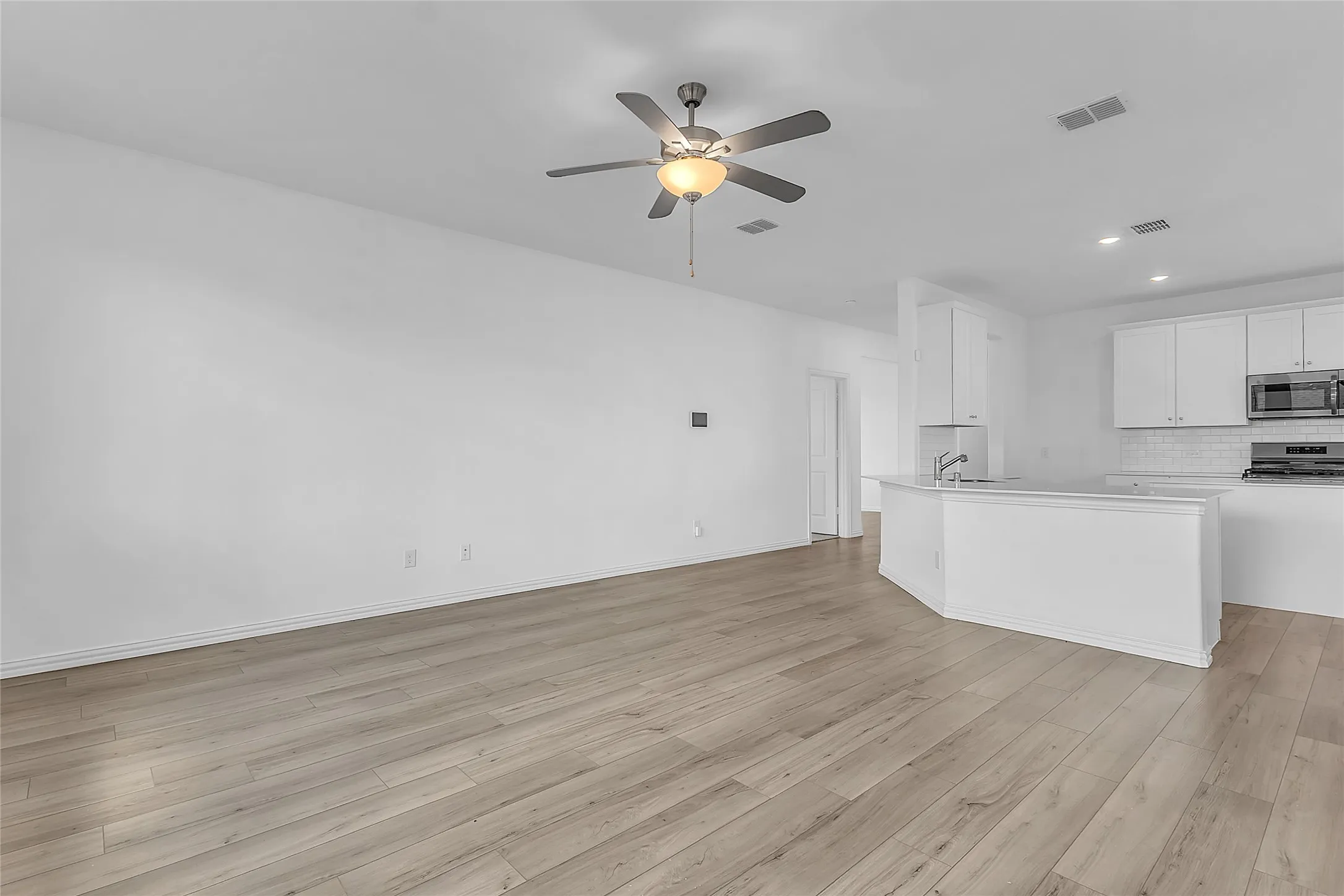 Living room with wood-look floors and a ceiling fan, open to the kitchen peninsula and white cabinetry