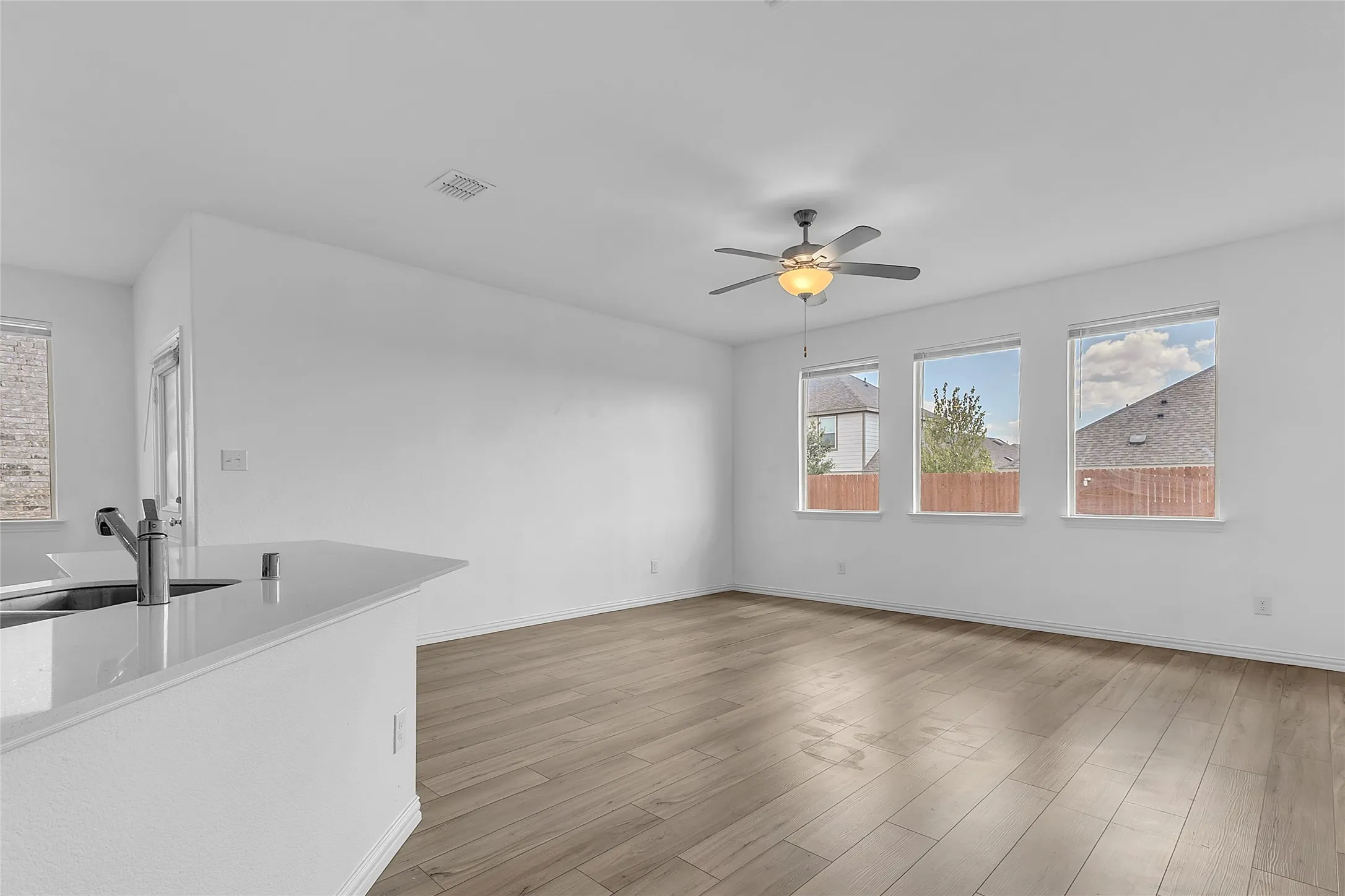 Living room with three windows and a ceiling fan with light, open to the kitchen's sink peninsula