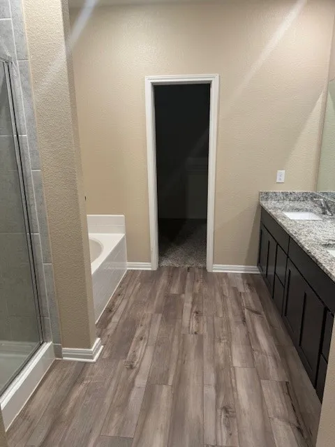 Wood-finish flooring throughout, featuring a built-in tub, separate glass-enclosed shower, and a double vanity with an integrated countertop