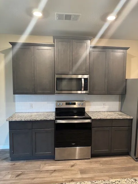 Kitchen featuring dark gray cabinetry, stainless steel appliances, granite countertops, white subway tile backsplash, and wood-finish flooring