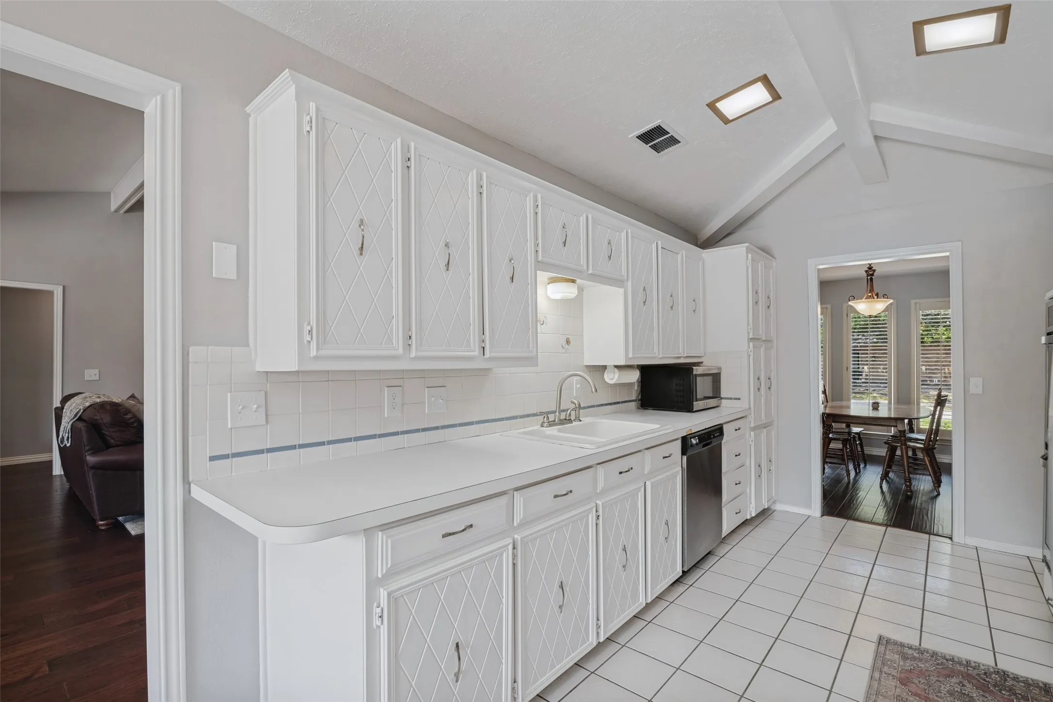White cabinetry with diamond pattern detailing, white tile backsplash, vaulted ceiling with exposed beams, stainless steel dishwasher, and tile flooring