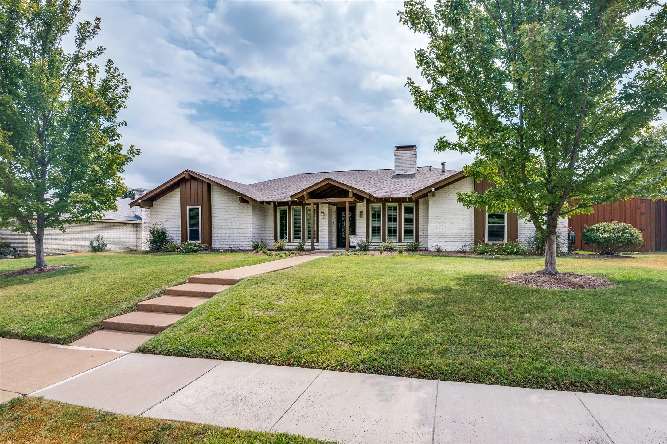 Contemporary ranch-style residence featuring a stucco exterior with wood-trimmed architectural accents, positioned on an expansive manicured lawn framed by mature shade trees