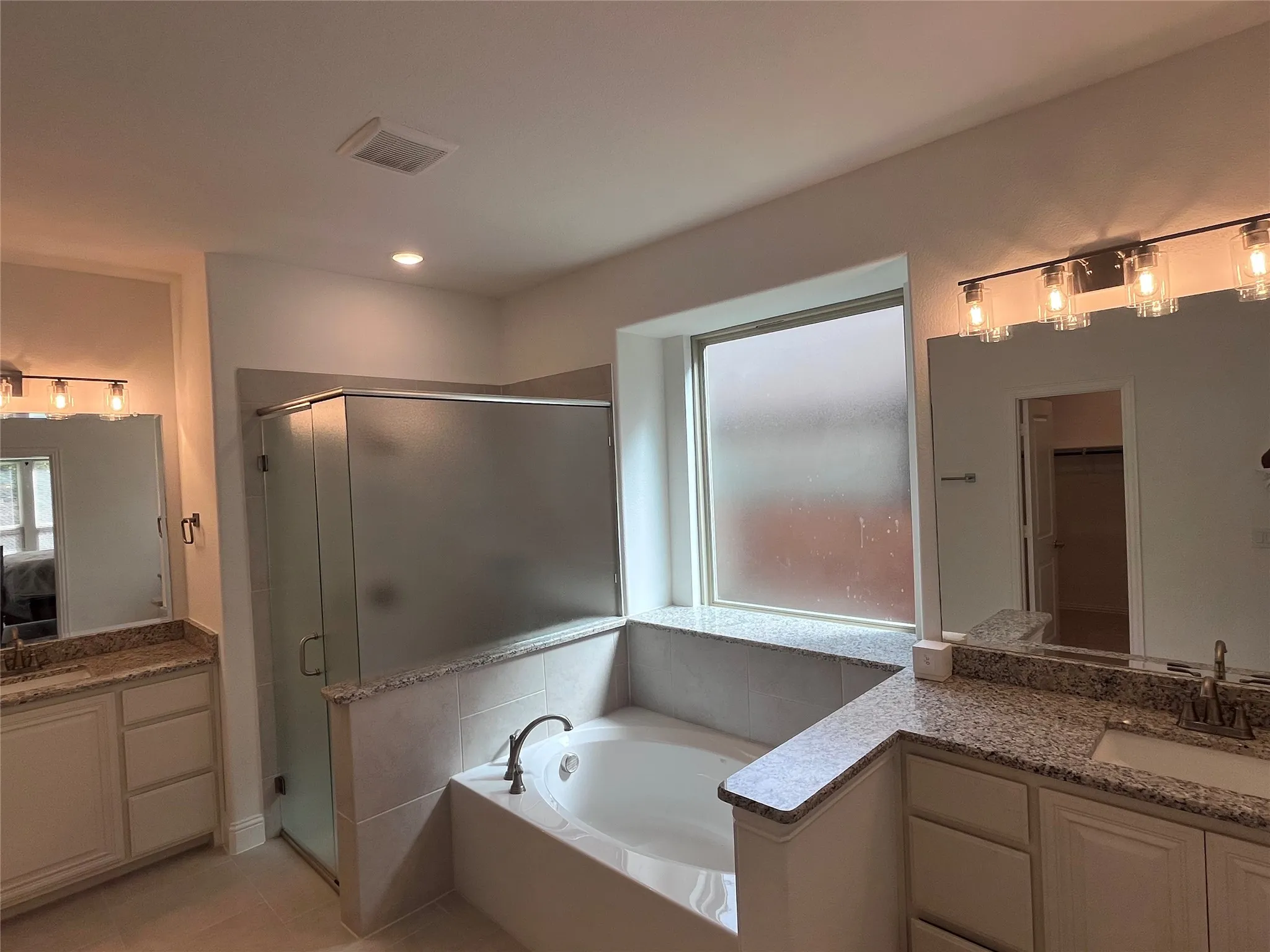 Dual vanities with granite countertops, separate shower with frosted glass enclosure, built-in soaking tub, and ample natural light from the large window