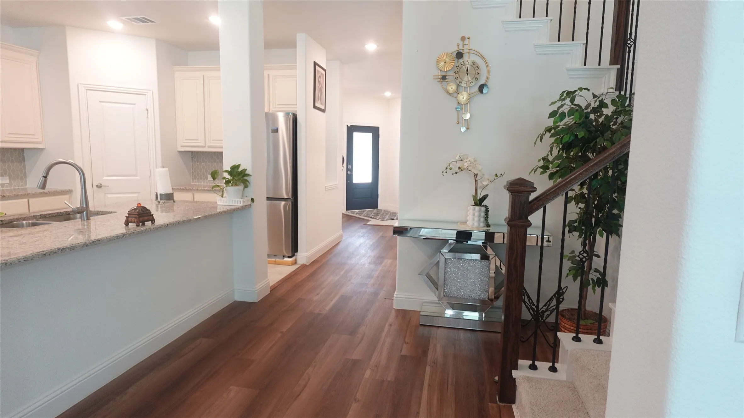 Open-concept living space with wood-finish flooring, a kitchen featuring light cabinetry and granite-style countertops, and a staircase with dark wood and wrought iron-style balusters