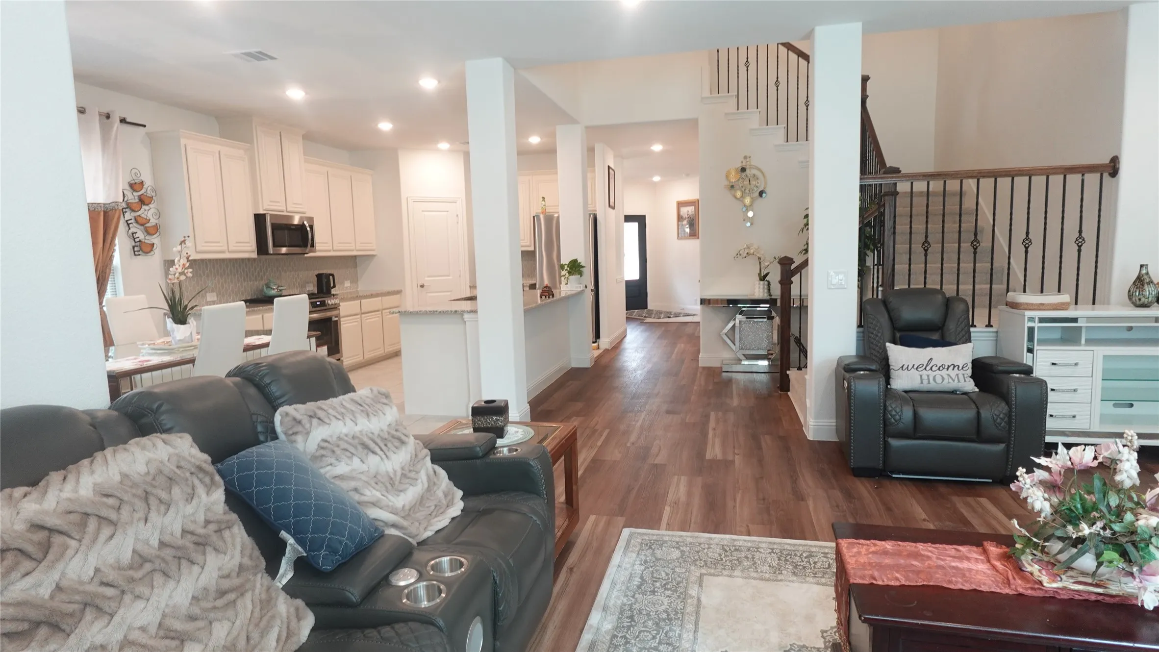 Open concept living space featuring wood-finish flooring, recessed lighting, a kitchen with white cabinetry and stainless steel appliances, and a staircase with decorative wrought iron spindles