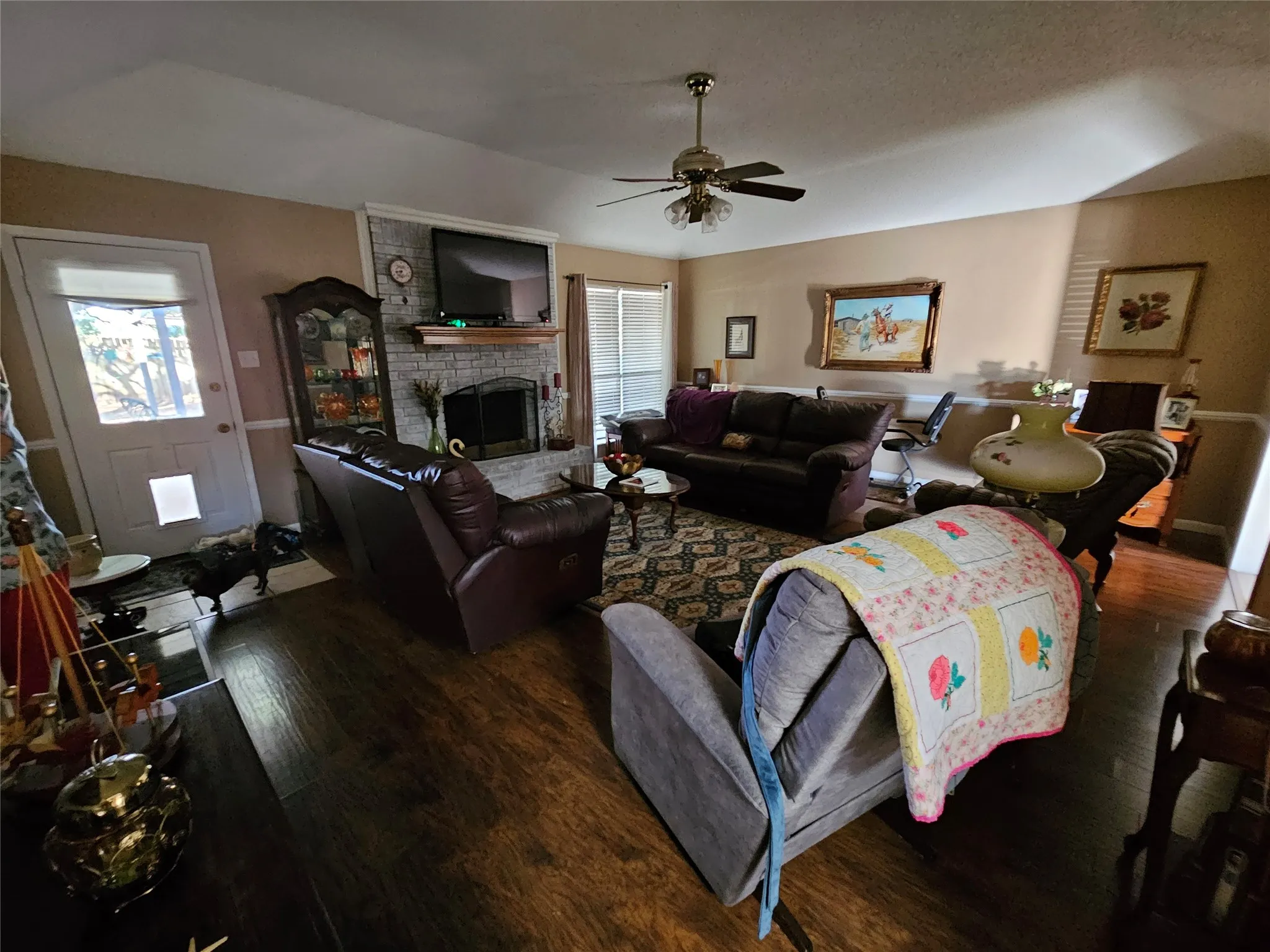 Wood-finish flooring, ceiling fan with integrated lighting, a white painted door with a window, and a brick fireplace with a mantel