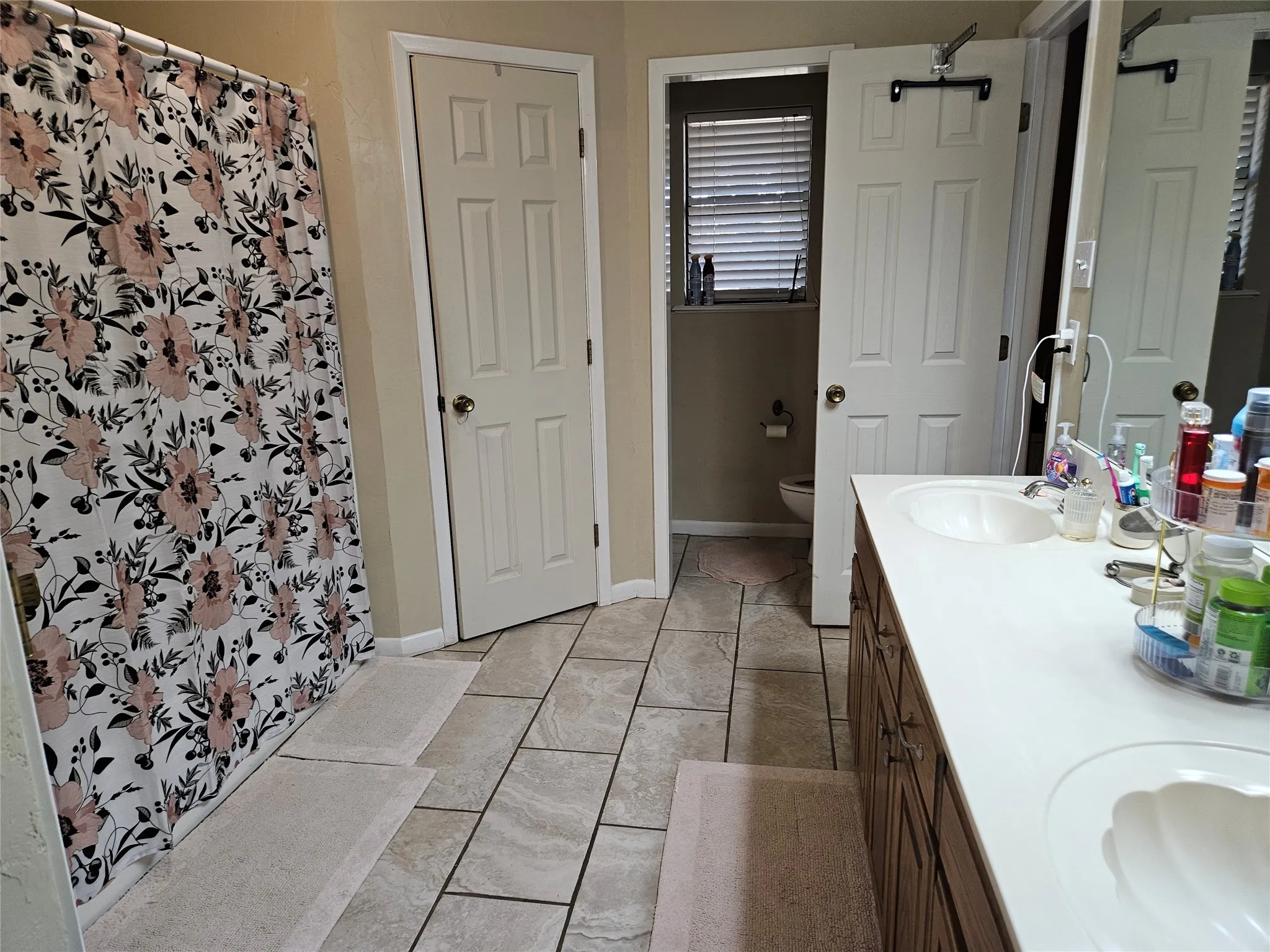 Double vanity with integrated sinks, wood-finish cabinetry, neutral tile flooring, and a window with blinds