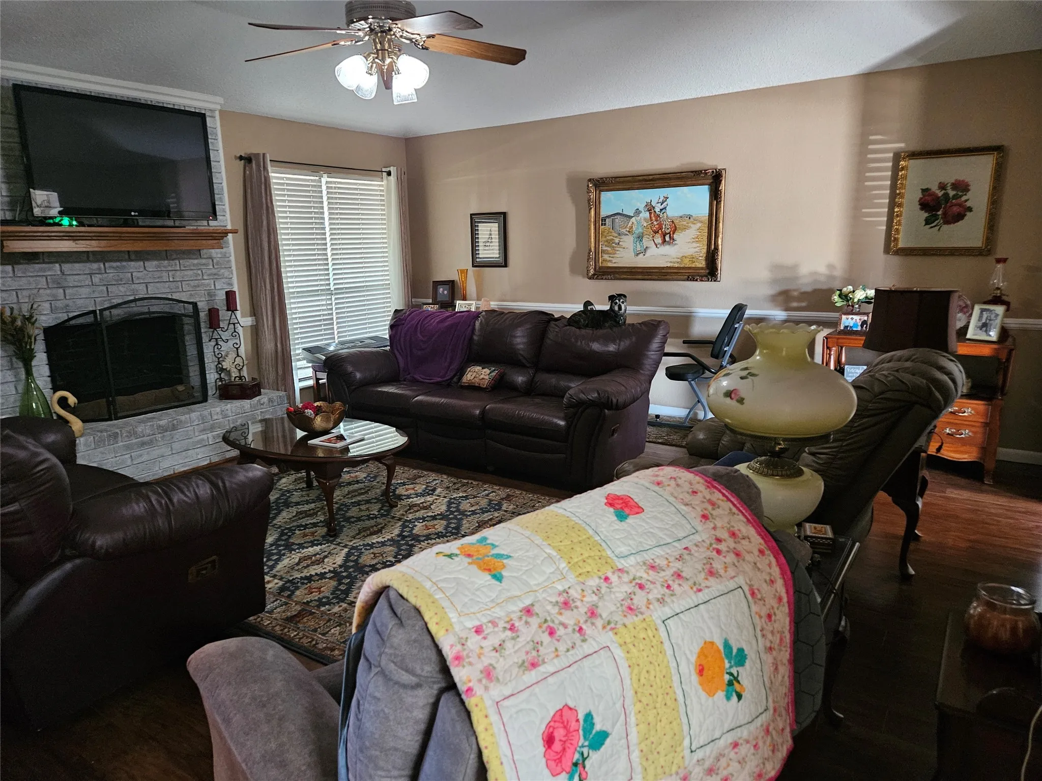 Fireplace with brick surround and wood mantle, wood-finish flooring, ceiling fan, and window with horizontal blinds