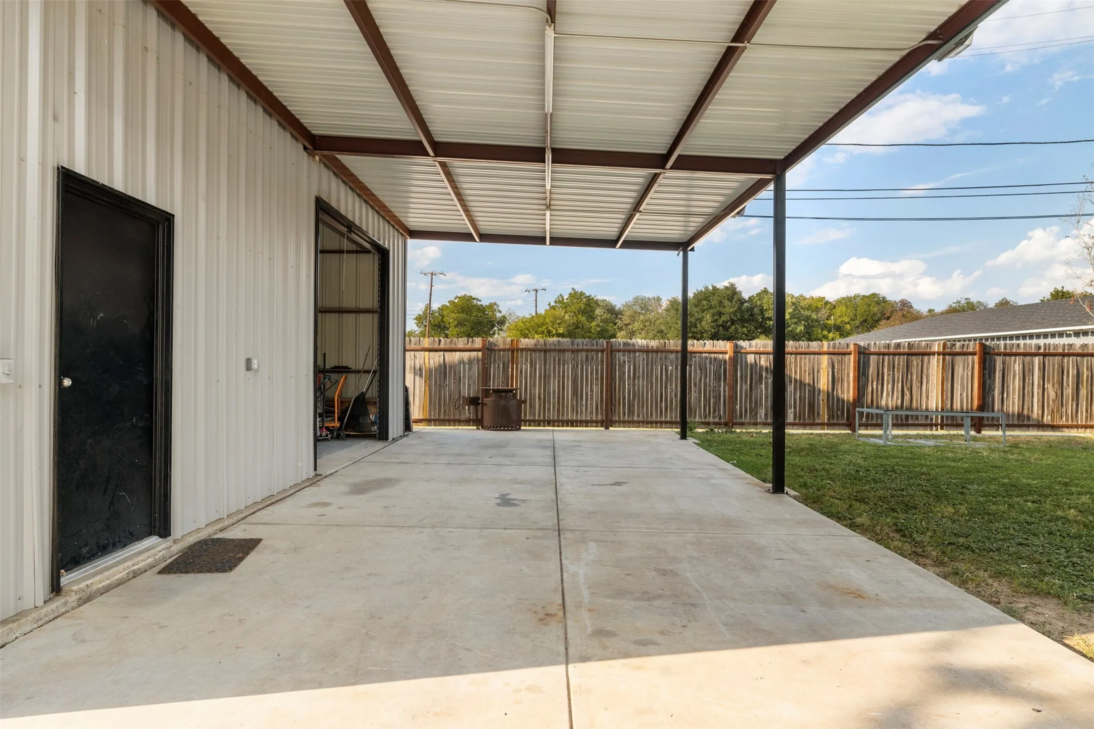 Covered concrete patio with a metal roof structure, vertical corrugated metal siding, and a wooden privacy fence