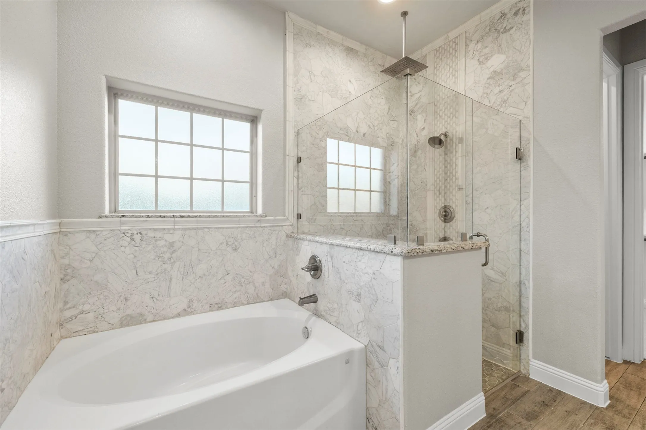 Corner soaking tub with marble-look surround, walk-in shower with frameless glass enclosure, rain shower head, and wood-look flooring