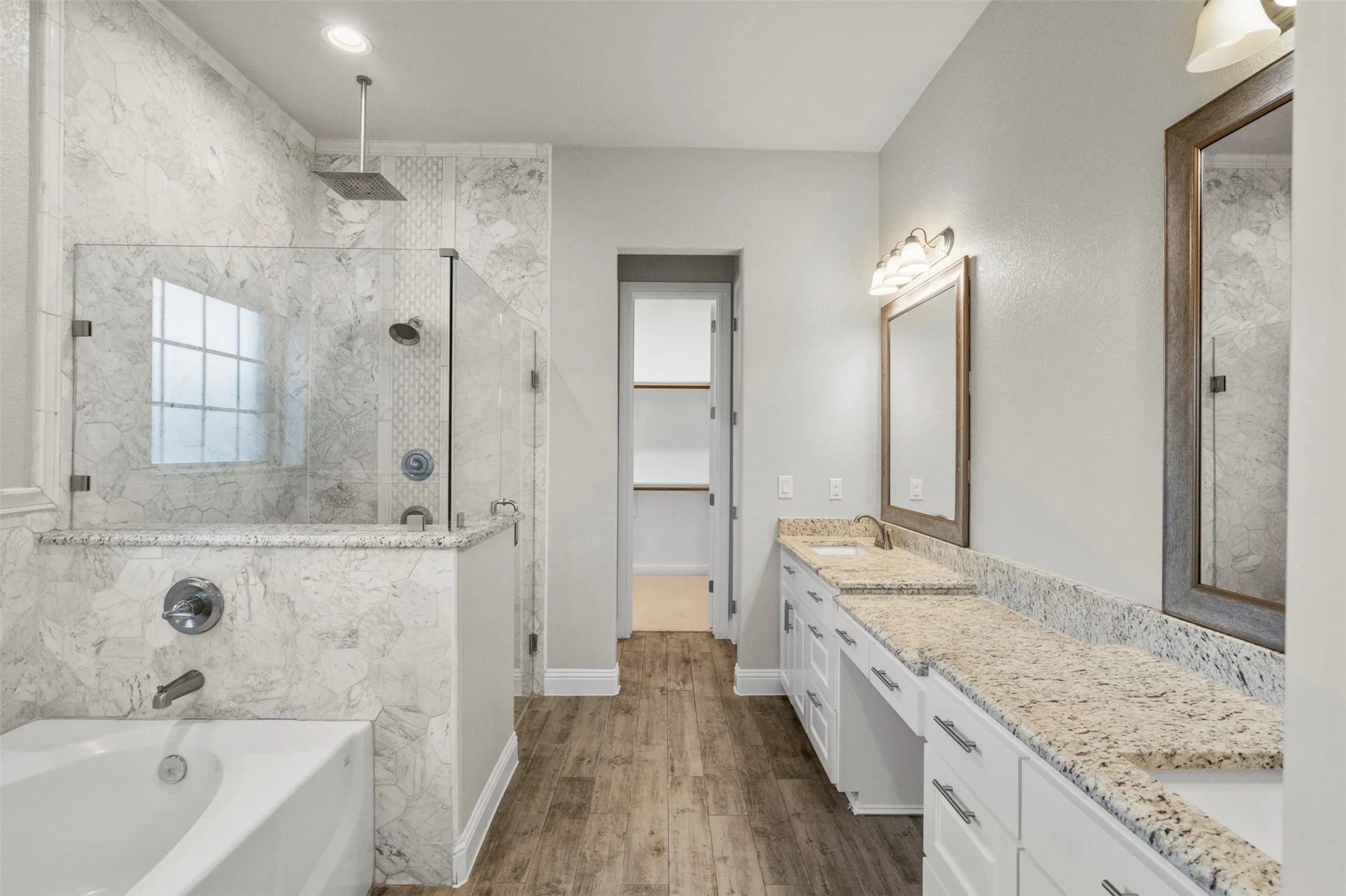 Spacious bathroom featuring a built-in bathtub, a walk-in shower with glass enclosure, dual vanity with granite countertops, and wood-finish flooring