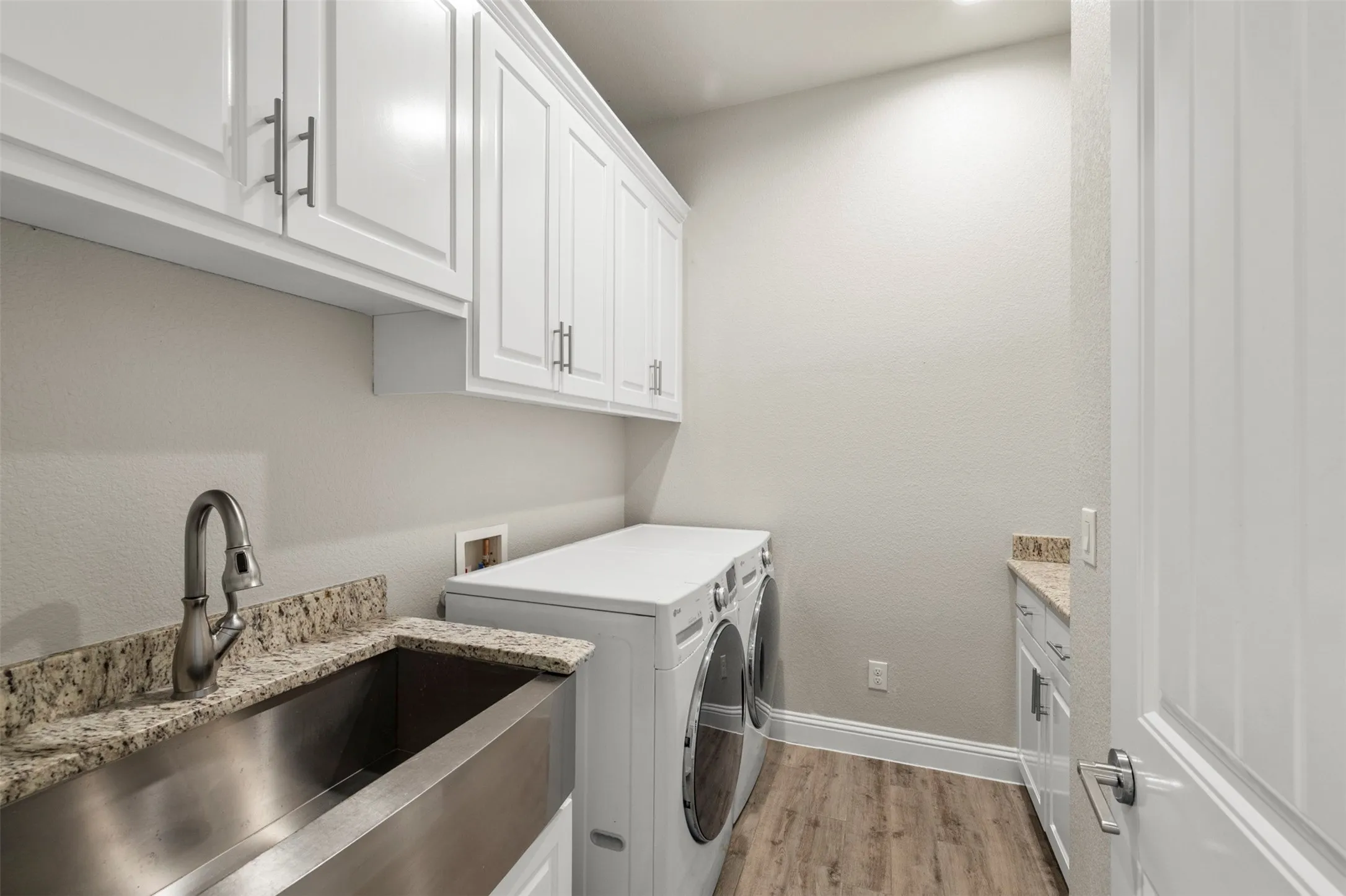 White shaker-style cabinetry with T-bar pulls, stainless steel farmhouse sink with gooseneck faucet, light-toned wood-finish flooring, and light-toned granite countertops