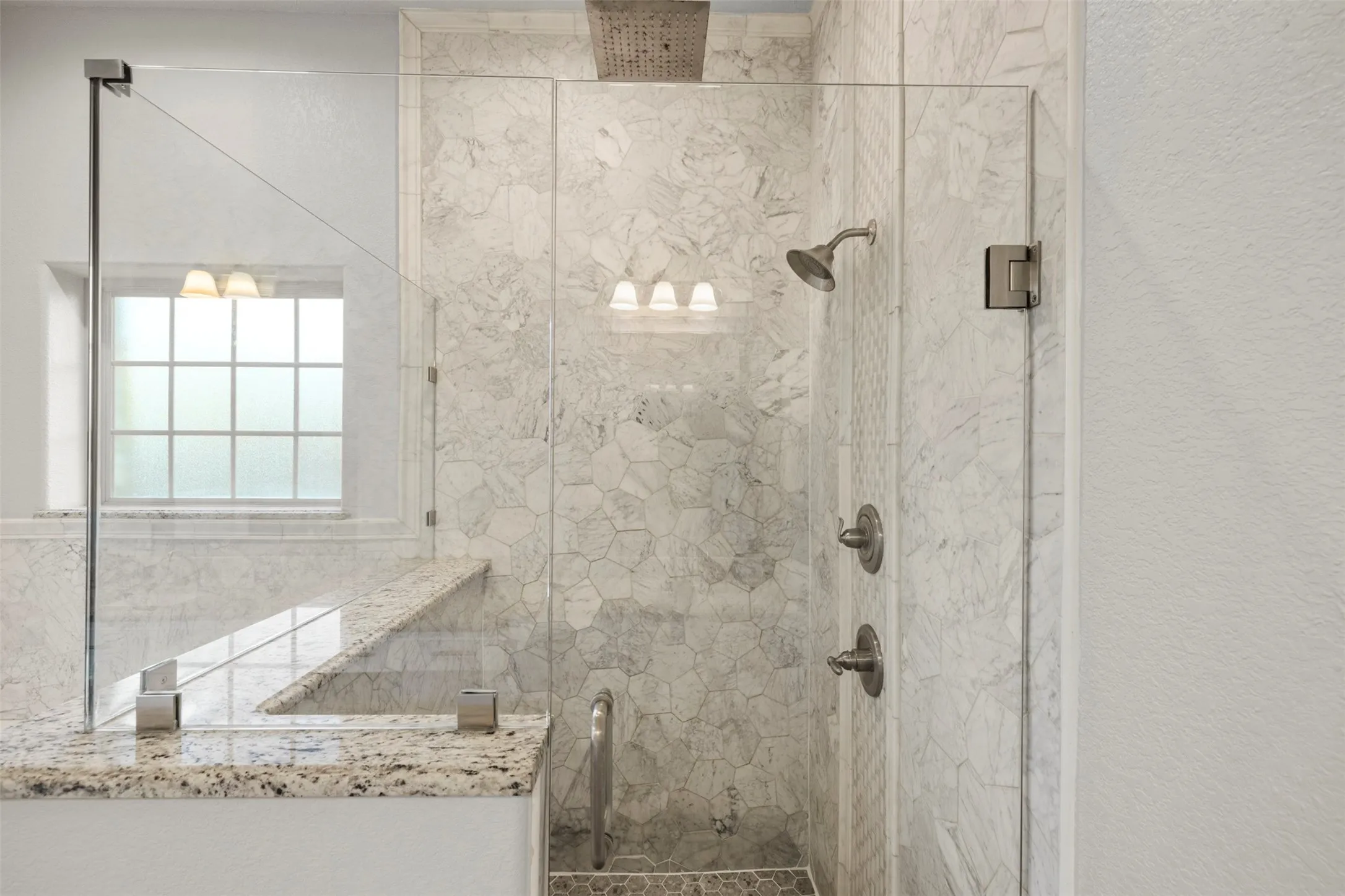 Frameless glass shower enclosure featuring a built-in bench with granite-look surface, large format tile walls, and a window with frosted glass