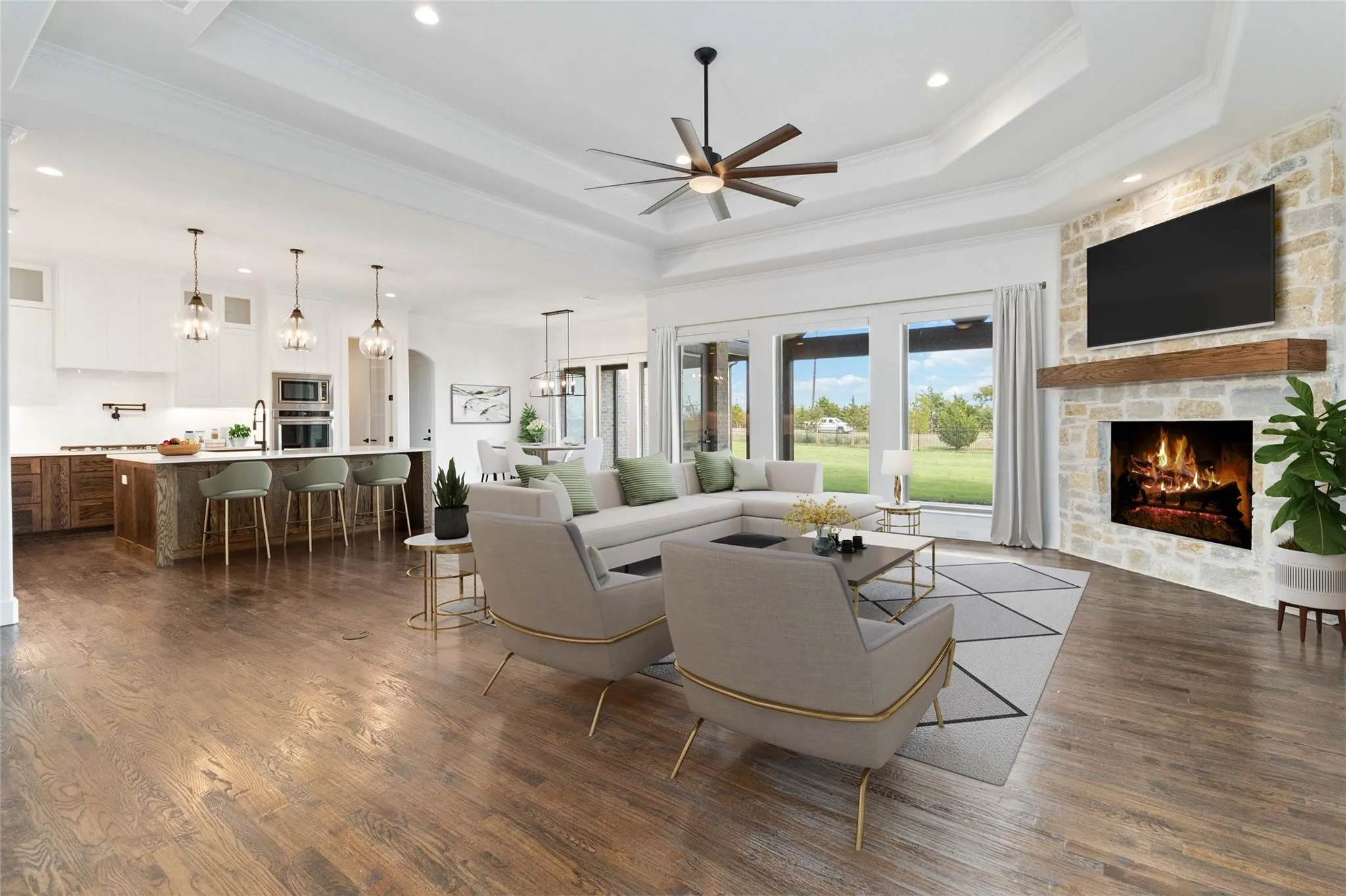 Tray ceiling with a ceiling fan, stone-clad fireplace with a wood mantel, wood-finish flooring, and a kitchen with an island featuring pendant lighting