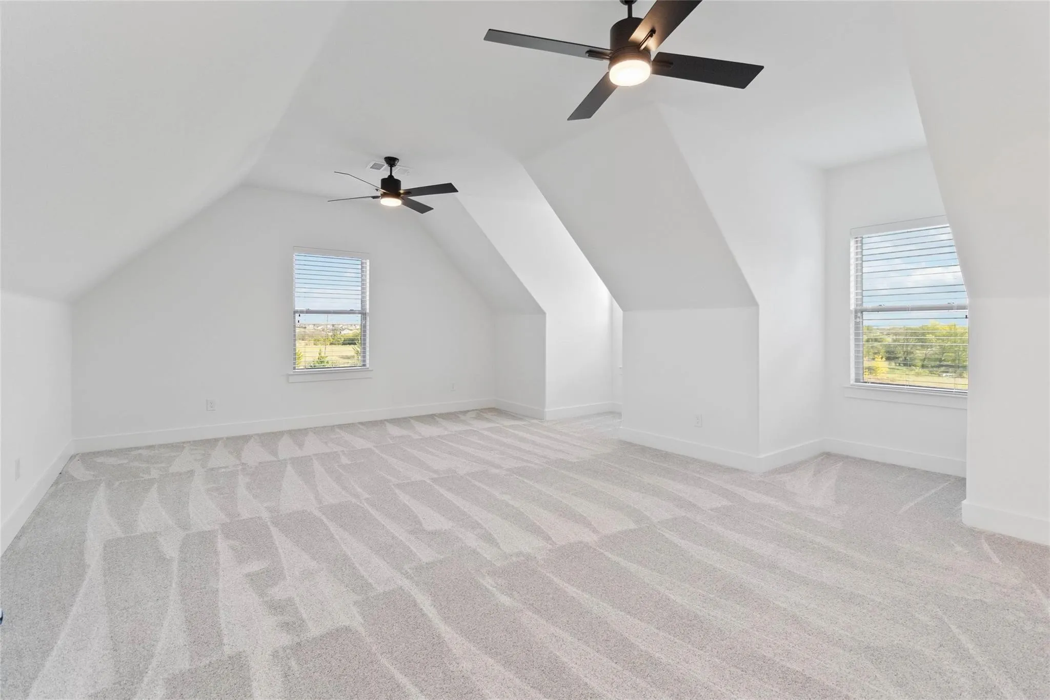 Spacious room featuring vaulted ceilings, light-toned patterned carpeting, multiple windows with blinds, and two ceiling fans