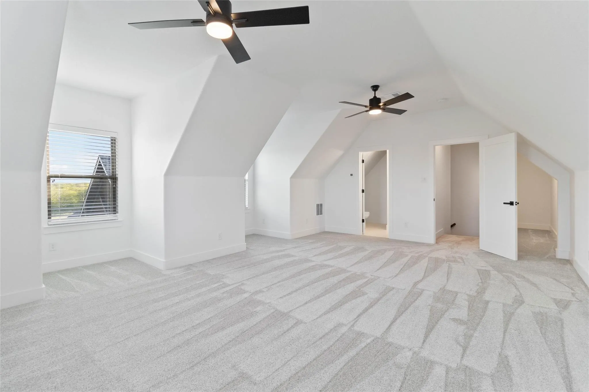Spacious room featuring vaulted ceilings, a window with blinds, two ceiling fans, and light-colored carpet