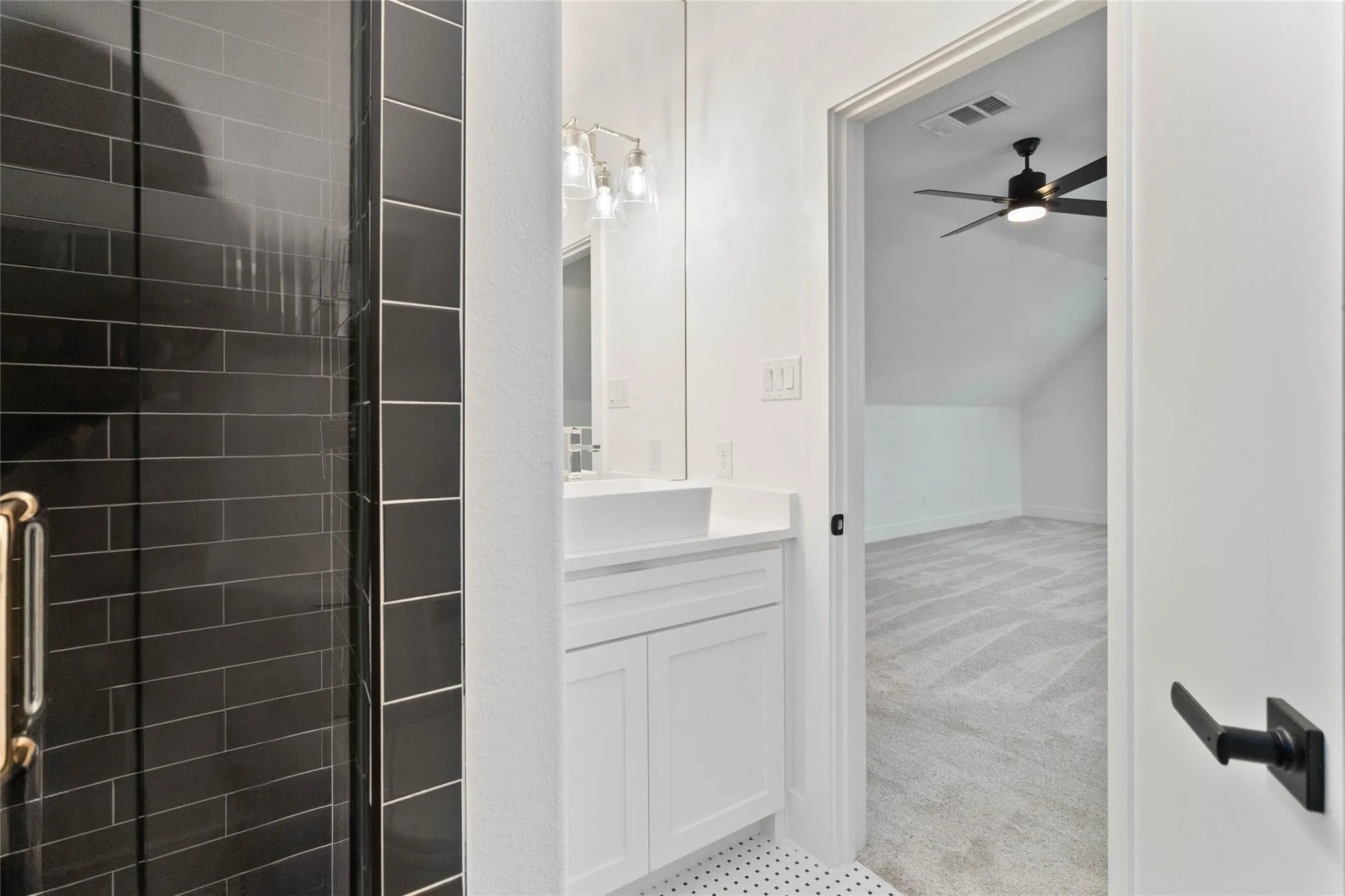 White vanity with integrated sink, modern light fixture, black subway tile shower, and textured carpet flooring