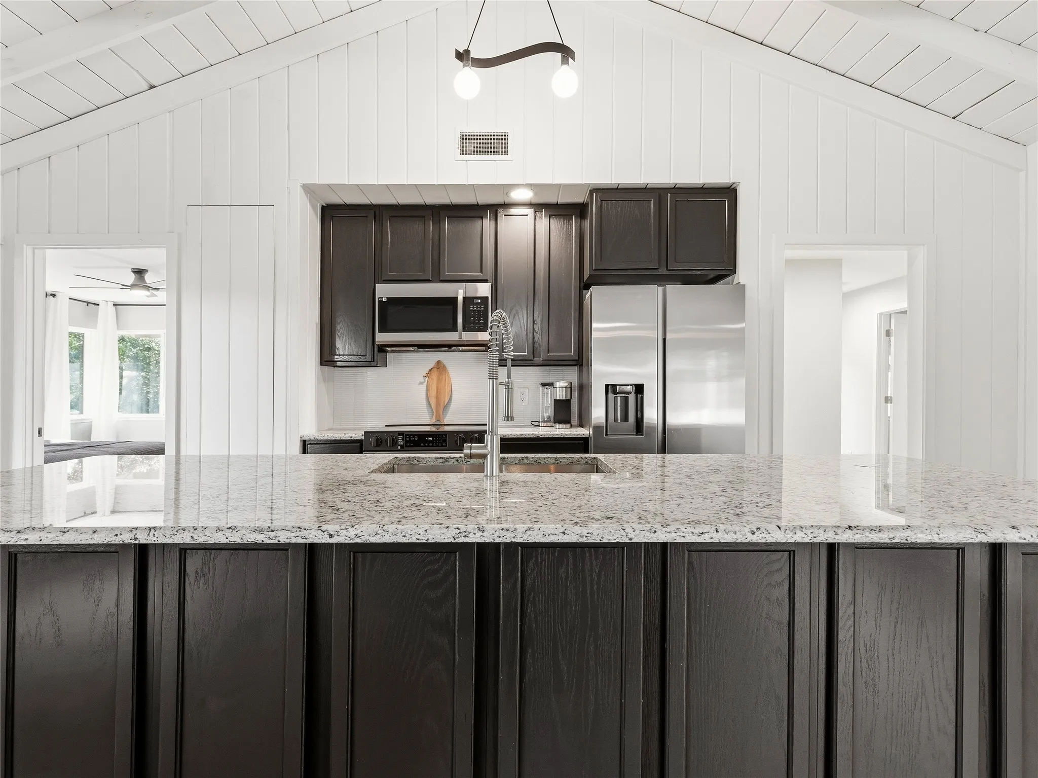 Vaulted ceiling with white plank paneling, dark wood-finish cabinetry, light speckled countertops, stainless steel appliances, and a kitchen island with an undermount sink
