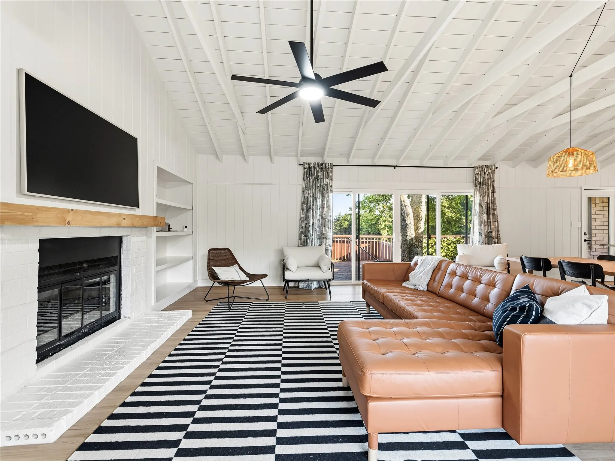 Vaulted ceiling with exposed beams, wood-finish flooring, white brick fireplace with timber mantel, built-in shelving, and sliding glass doors