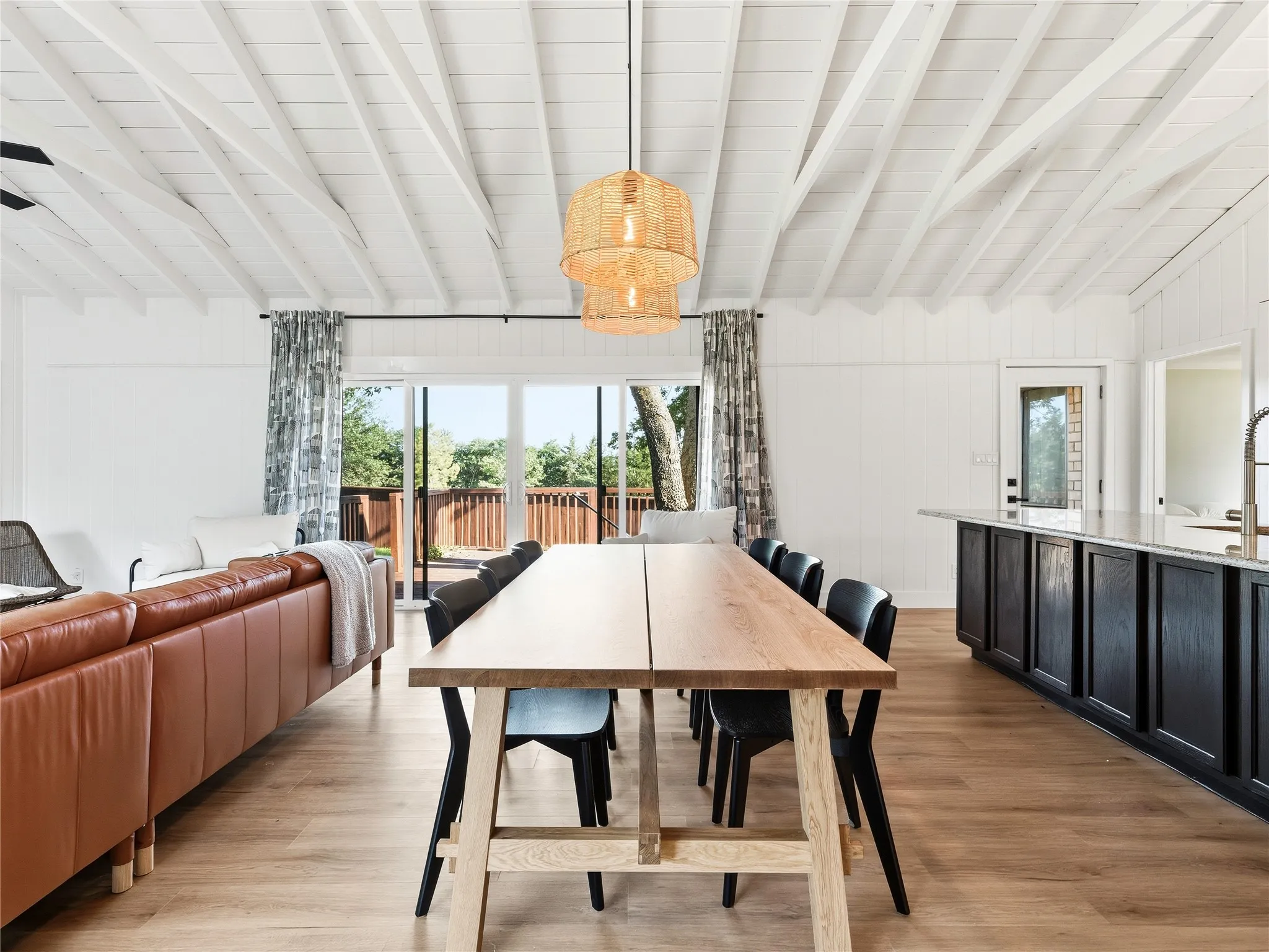 Open-concept living area with exposed beam ceiling, wood-finish flooring, sliding glass doors to outdoor deck, and kitchen island with built-in cabinetry and stone countertop