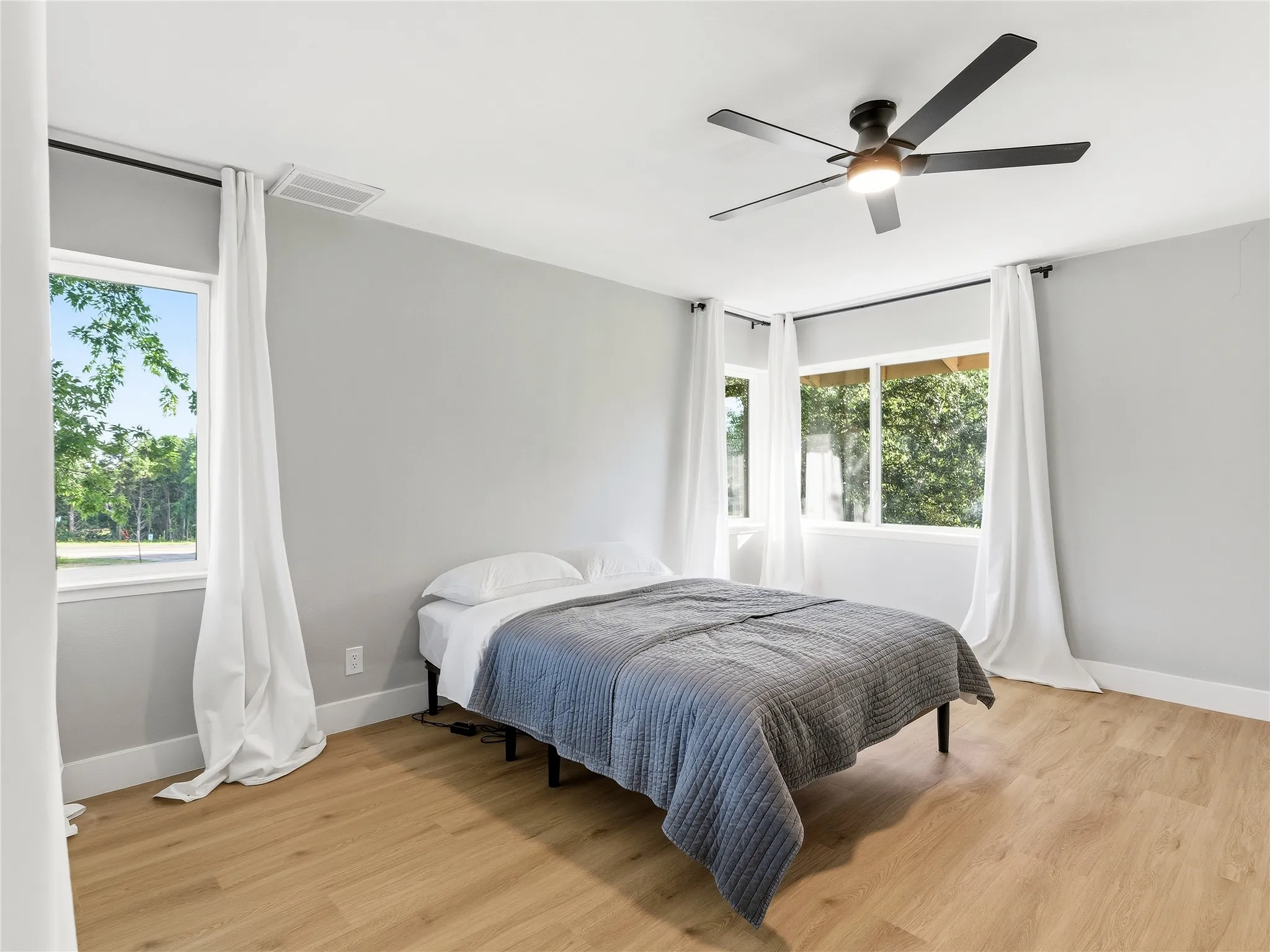 Spacious room featuring wood-finish flooring, multiple windows with white trim, a ceiling fan with integrated lighting, and light gray wall paint