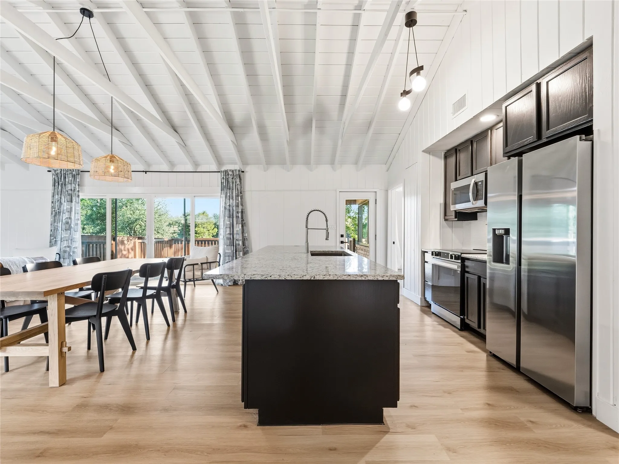Vaulted ceiling with exposed beams, wood-finish flooring, stainless steel appliances, kitchen island with integrated sink, and large sliding glass doors