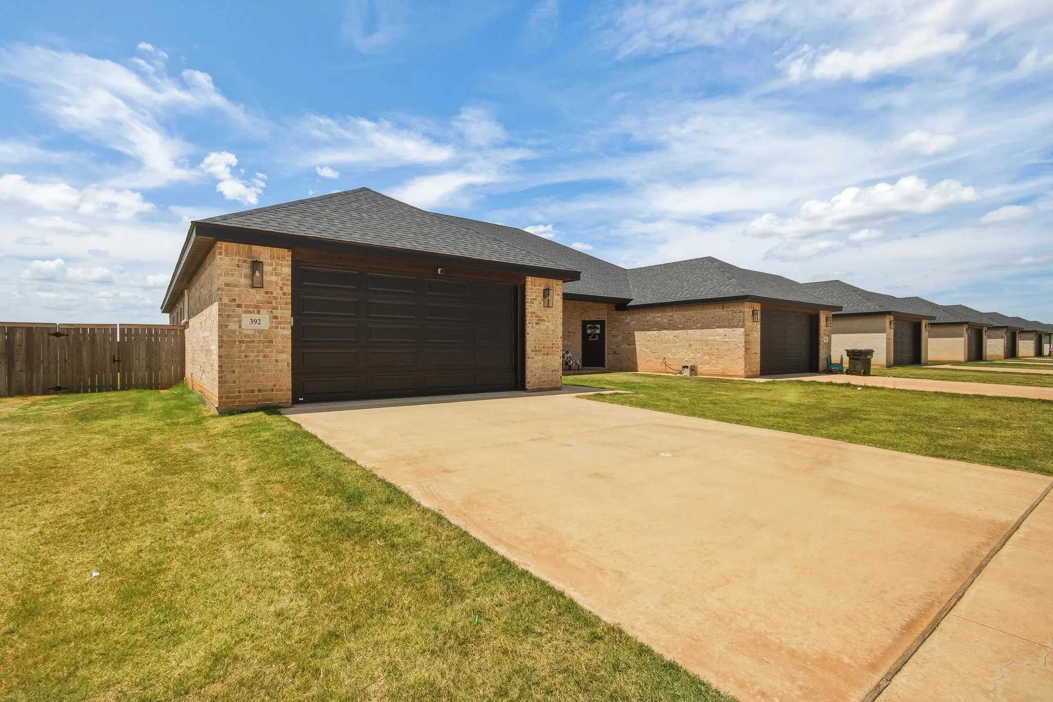 Brick exterior with dark asphalt shingle roof, attached garage with dark panel door, concrete driveway, and a wood privacy fence