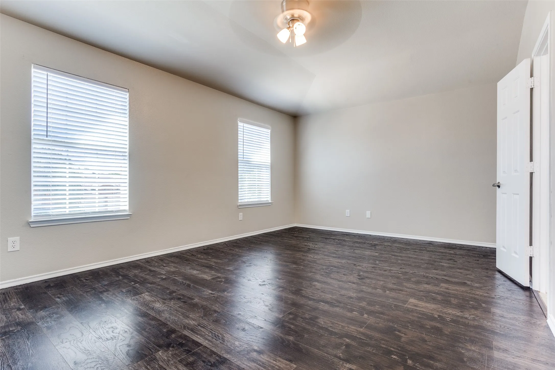 Spacious bedroom room featuring wood-finish flooring, two windows with blinds, a ceiling fan, and white trim