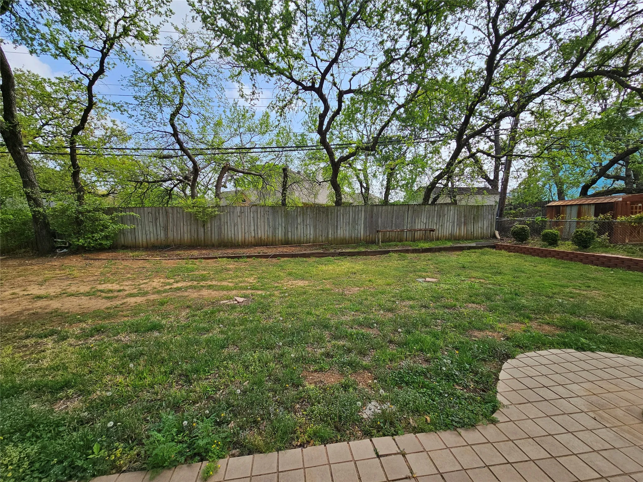 Grassed yard with mature trees, perimeter fencing, brick-lined planting beds, and a paver patio
