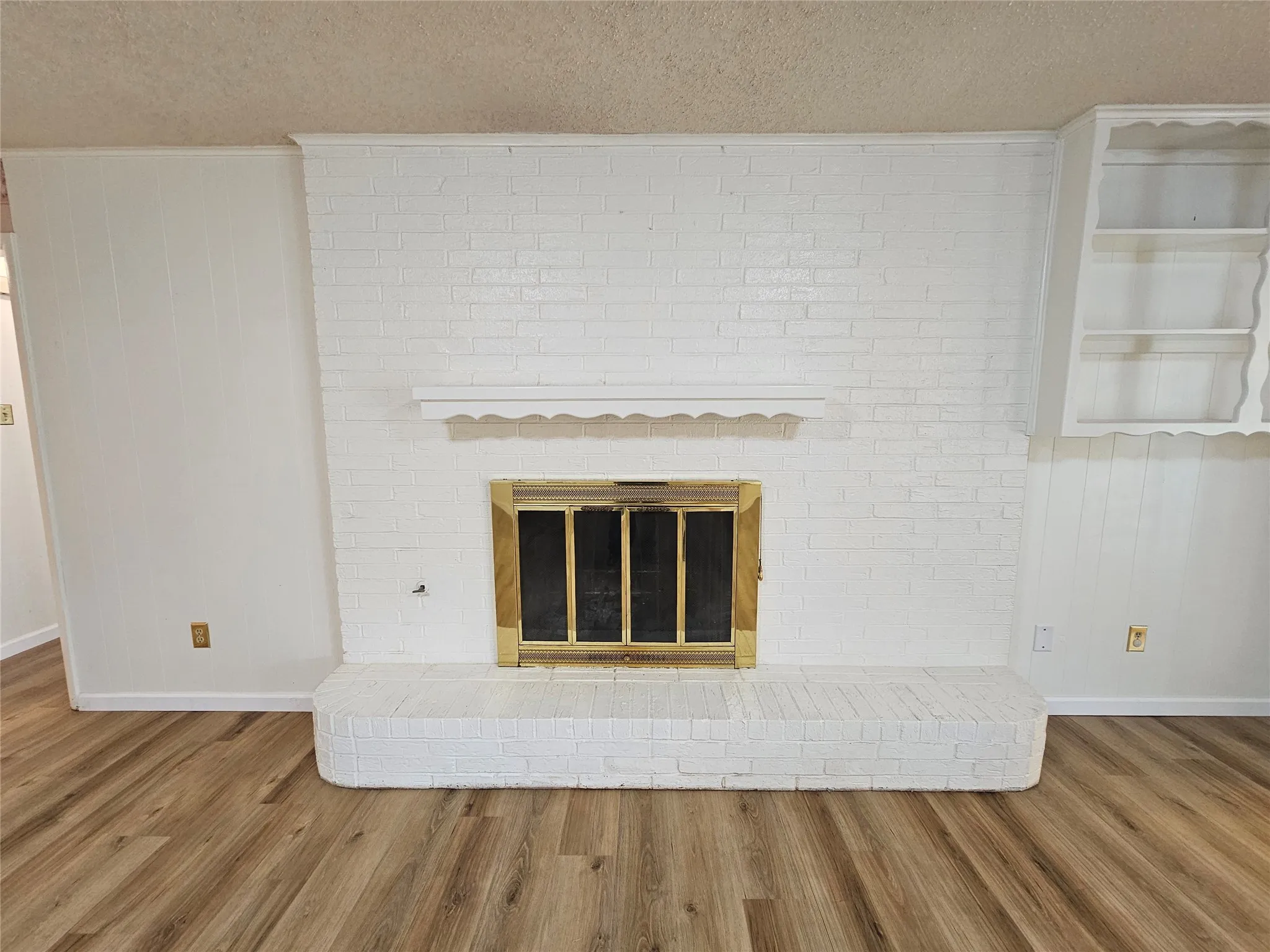 Brick fireplace with brass firebox, built-in shelving, wood-finish flooring, and painted paneling