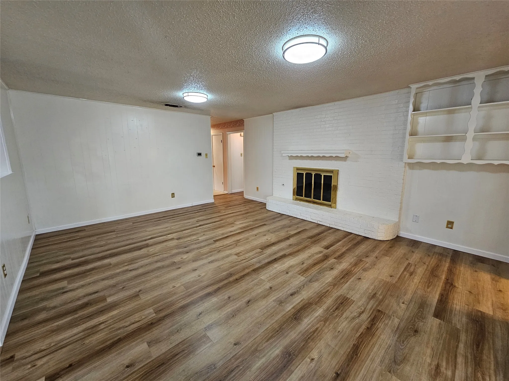 Wood-finish flooring, white painted brick fireplace with brass insert, built-in shelving, and two flush-mount ceiling lights