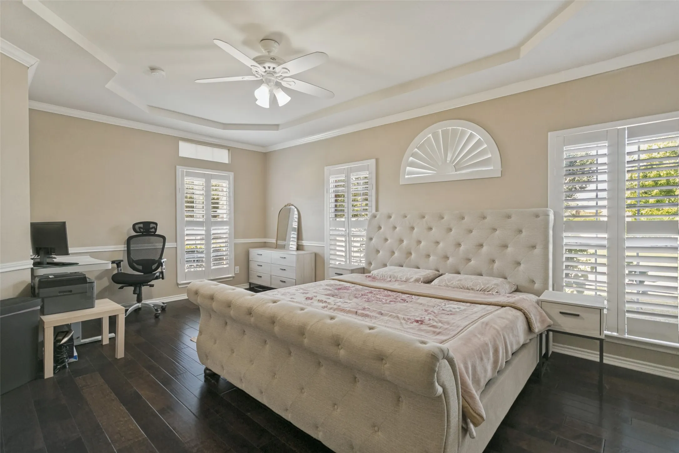 Tray ceiling with crown molding, dark wood-finish flooring, white plantation shutters, and a ceiling fan with integrated lighting
