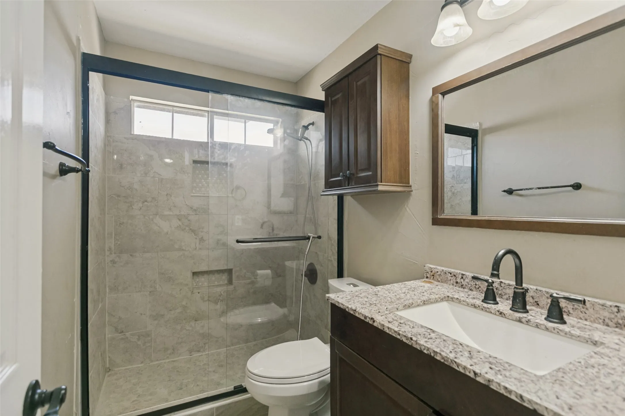 Spa-inspired bathroom featuring a walk-in shower with glass enclosure and tile surround, dual vanity with granite countertop and undermount sink, matte black fixtures, framed mirror, and recessed ceiling lighting