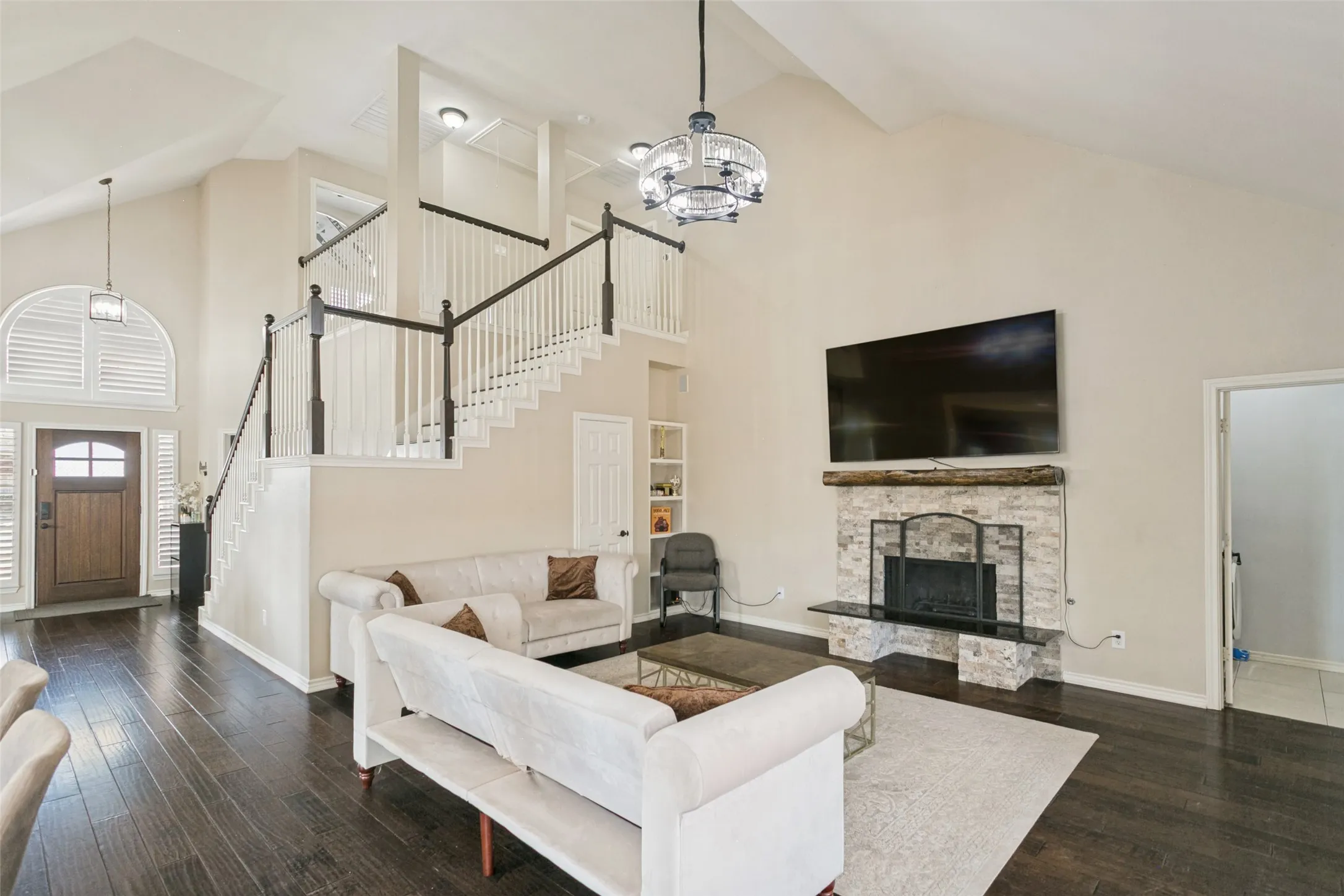 Vaulted ceiling with a decorative fan fixture, dark wood-finish flooring, stone-clad fireplace with mantel, staircase with dark wood handrails and white balusters, and a front entry door with an arched transom