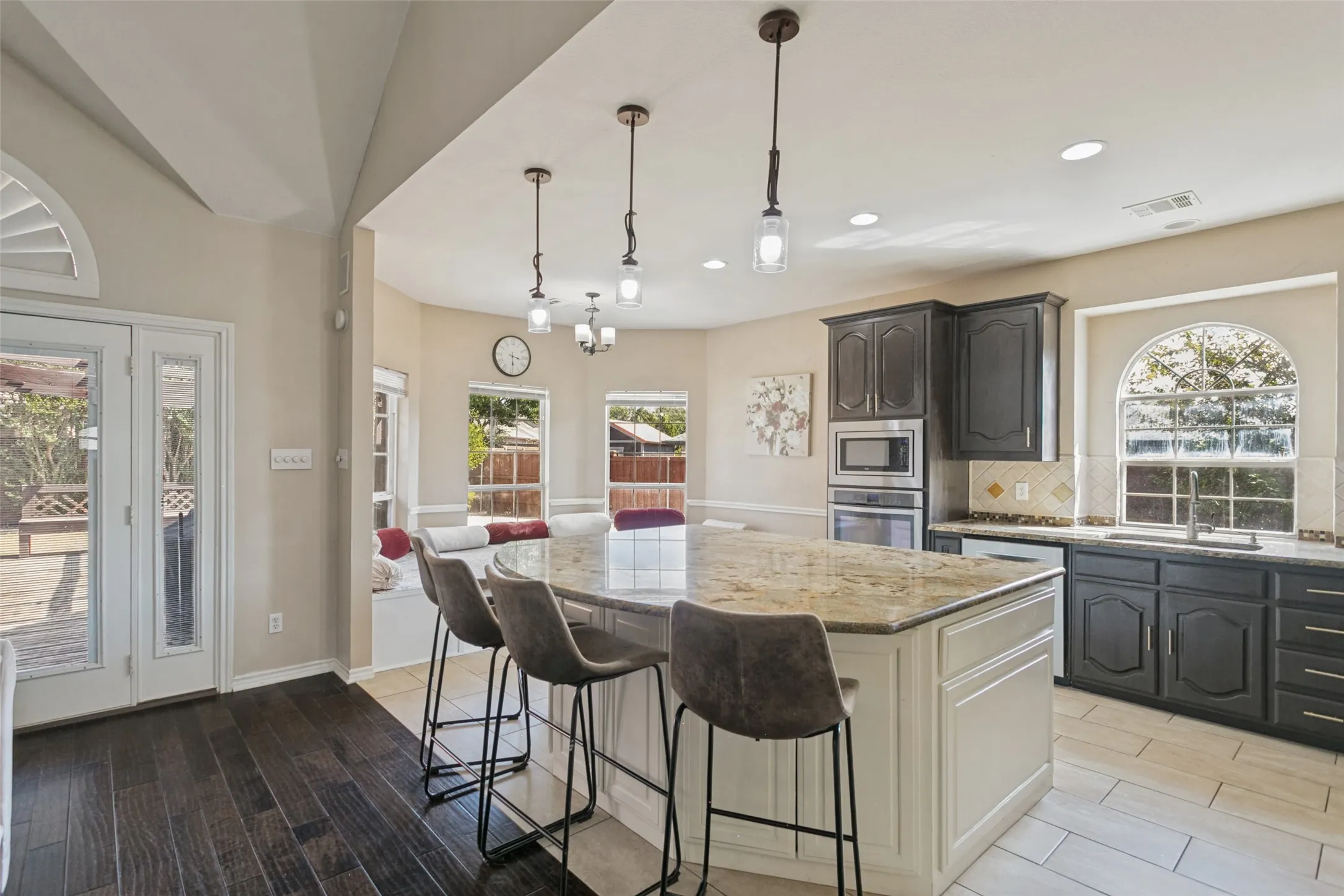 Gourmet kitchen featuring a large center island with wood-finish countertops and seating for multiple guests, dark cabinetry with contrasting cream-painted perimeter cabinets, stainless steel built-in appliances, pendant lighting, and abundant natural light from multiple windows