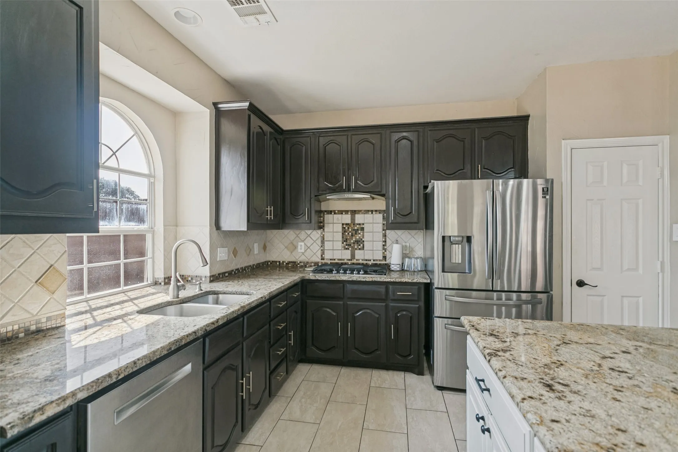 Kitchen featuring dark wood cabinetry, light-toned granite countertops, a stainless steel refrigerator, and a tile backsplash