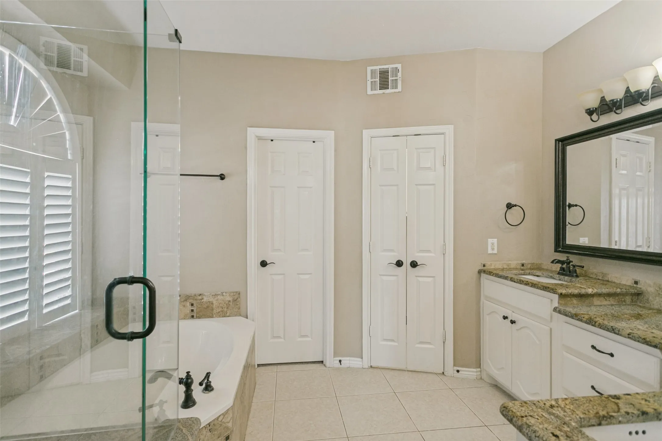 Glass-enclosed shower with a deep soaking tub, plantation shutter window, dual vanity with granite-style countertops, and tile flooring