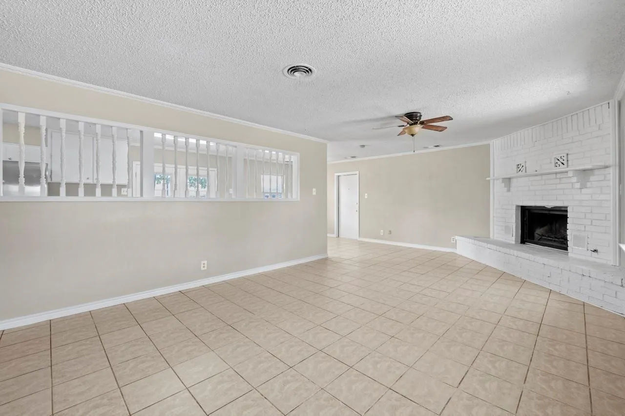Spacious room featuring neutral-toned tile flooring, a painted brick fireplace, a ceiling fan, and a decorative interior half-wall with spindles