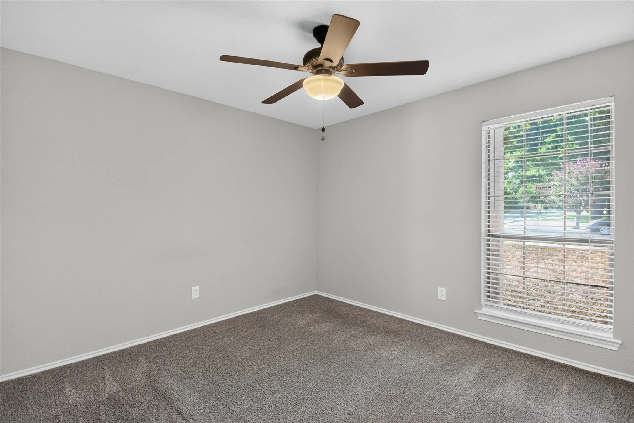 Bright corner bedroom with neutral wall finishes, plush carpeting, ceiling fan with integrated light fixture, and large window with plantation shutters providing natural light and privacy