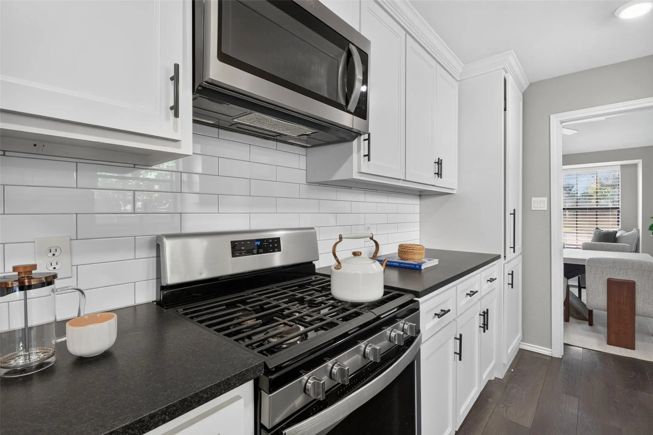 White shaker cabinetry, dark countertops, white subway tile backsplash, stainless steel microwave, and wood-finish flooring