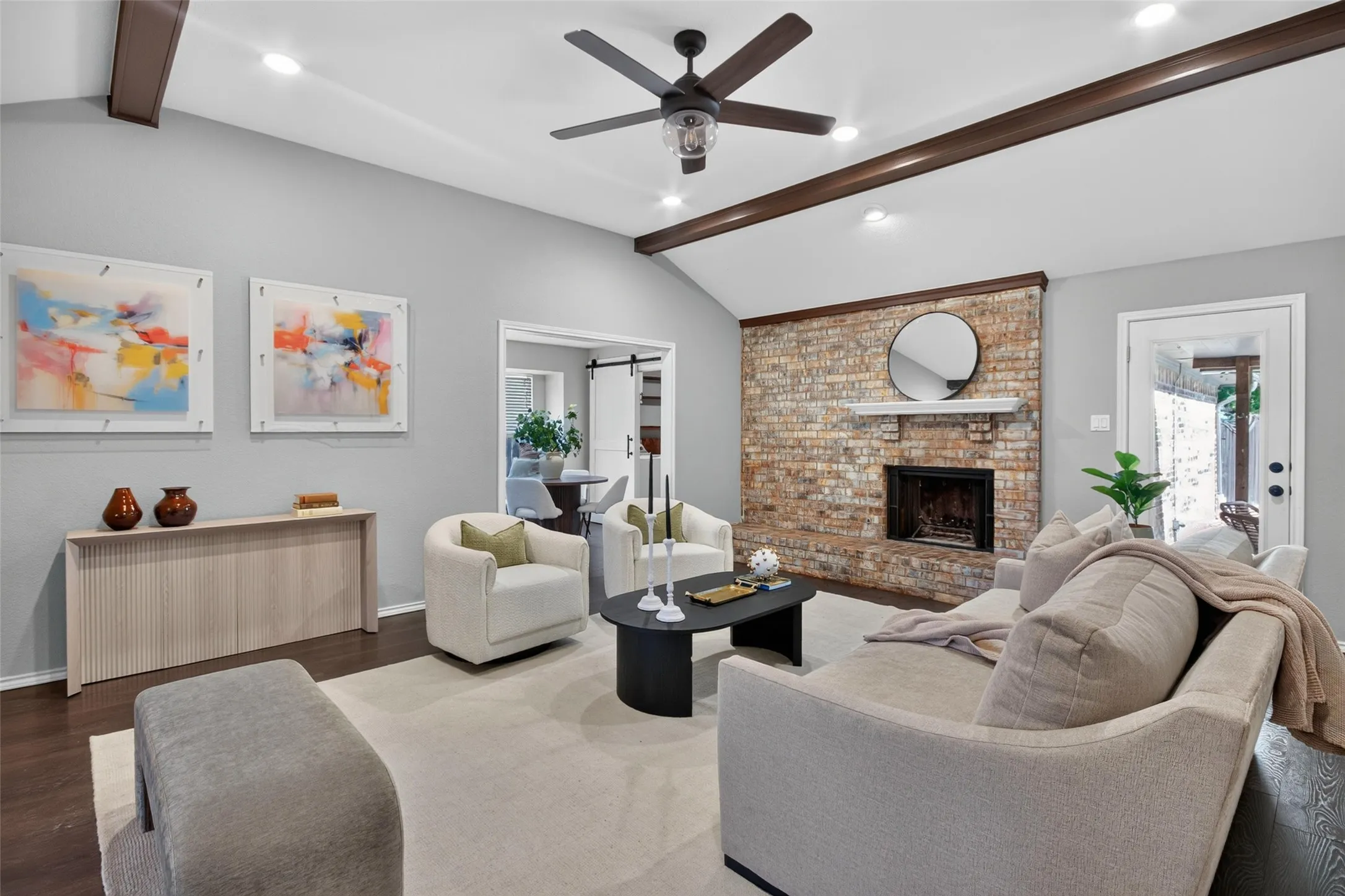 Vaulted ceiling with exposed wood beams, brick fireplace with mantel, dark wood-finish flooring, recessed lighting, and a ceiling fan