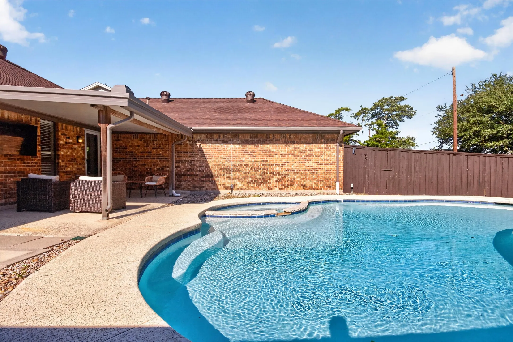 Curved swimming pool with an integrated spa, complemented by a covered patio, brick exterior, and a privacy fence