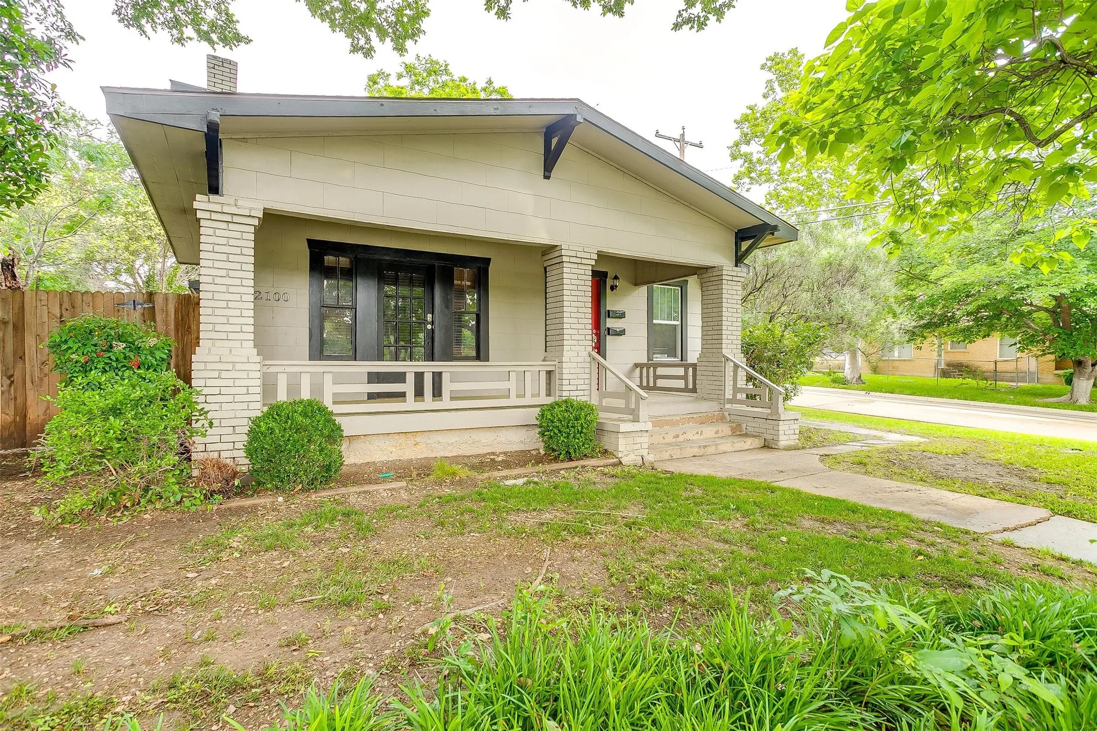 Front exterior featuring a covered porch with brick columns, a wood fence, and mature trees