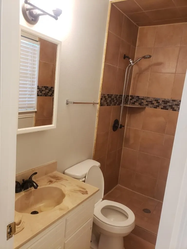 Newly updated bathroom featuring ceramic tile walls with decorative mosaic accent band, wood-finish vanity with integrated sink, white porcelain toilet, and walk-in shower with dual showerheads and tile surround