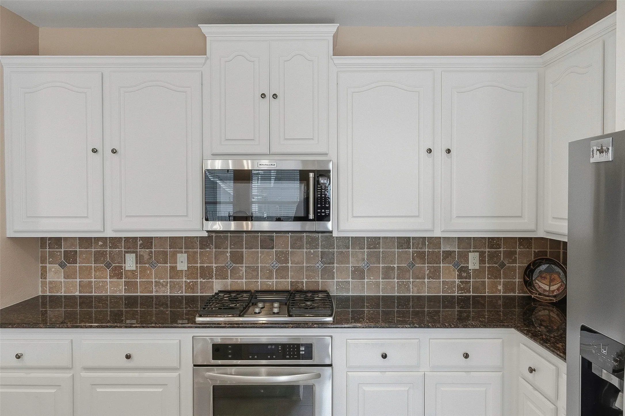 White cabinetry with crown molding, patterned tile backsplash, dark-toned countertops, built-in stainless steel microwave, and stainless steel oven with gas cooktop