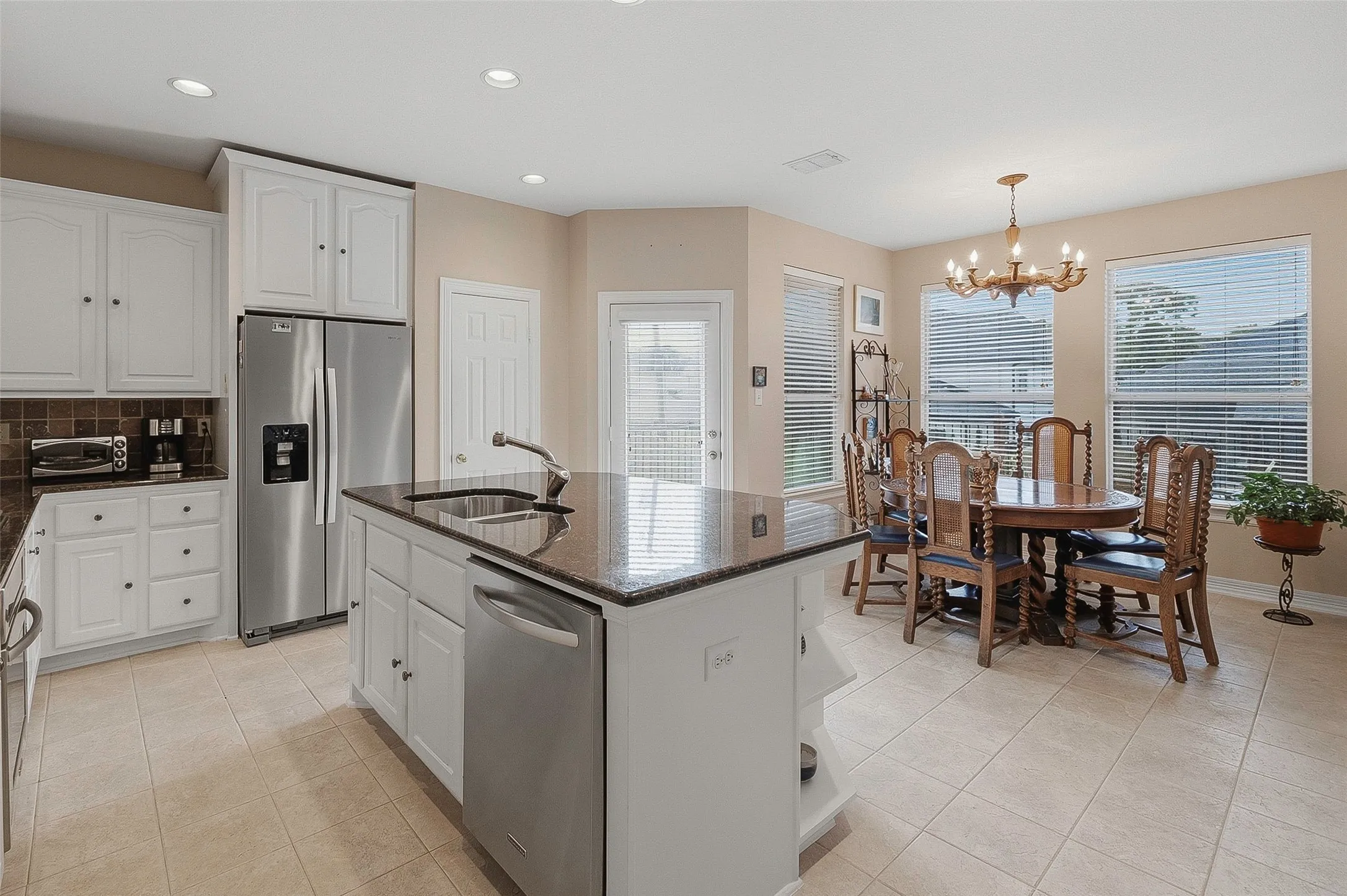 Tile flooring, kitchen island with integrated sink, white cabinetry, stainless steel appliances, and a dining area with multiple windows