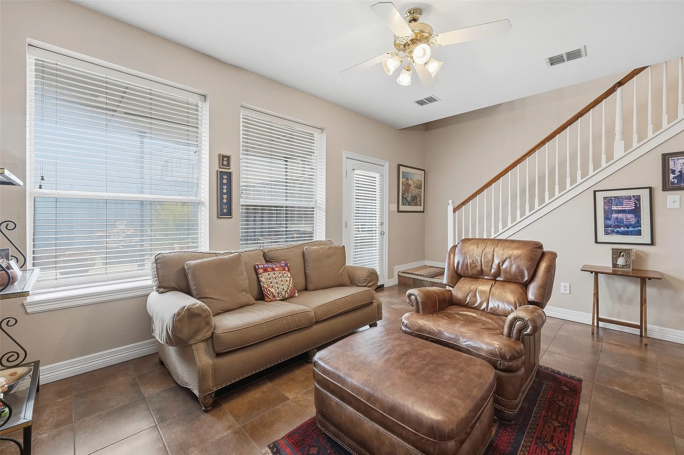 Living area featuring large windows with blinds, a ceiling fan with integrated lighting, a staircase with wood-finish handrail and white balusters, and tile flooring