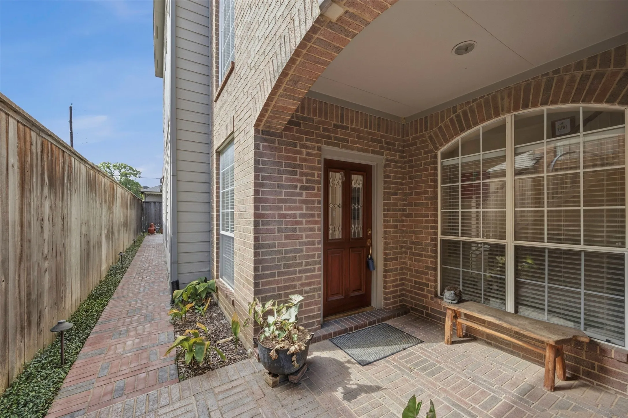 Brick exterior with covered patio featuring herringbone paver flooring, large multi-pane windows, wood-finish entry door, and mature landscaping with potted plants along the perimeter
