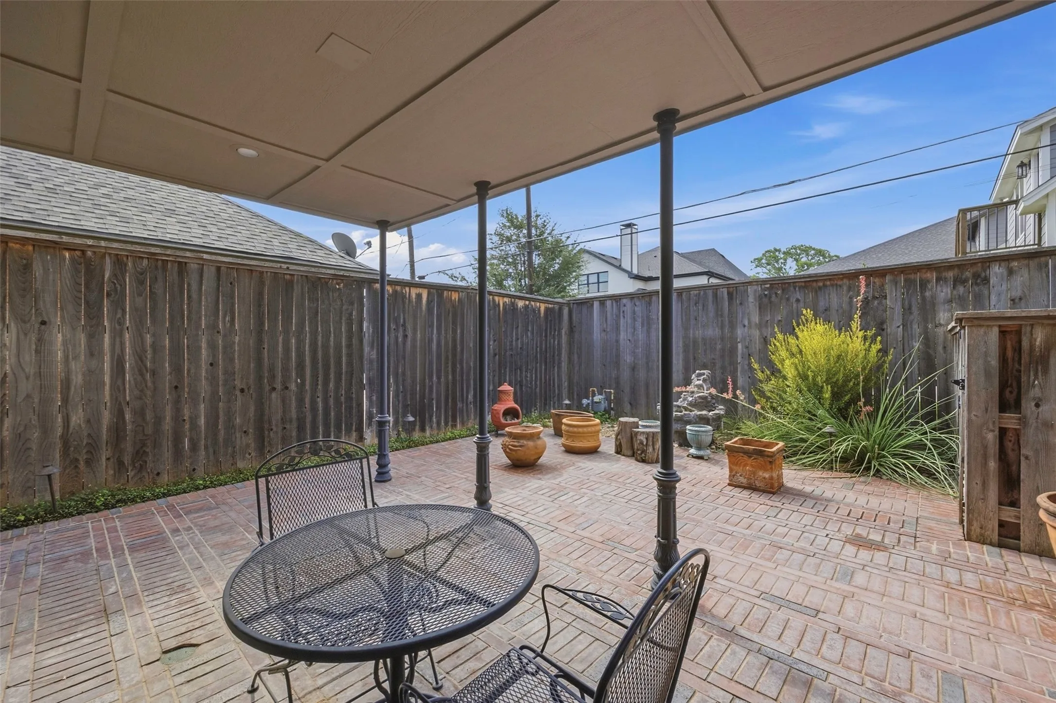 Covered patio with brick paver flooring, supported by dark decorative columns, and featuring a privacy fence
